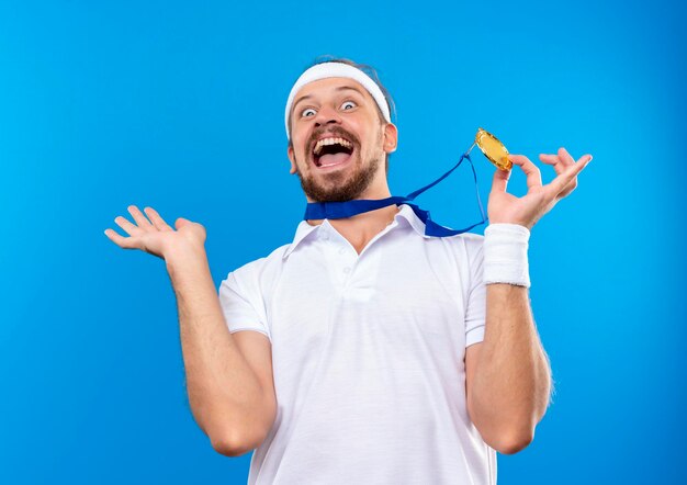 Joyful young handsome sporty man wearing headband and wristbands and medal around neck holding medal and showing empty hand isolated on blue wall