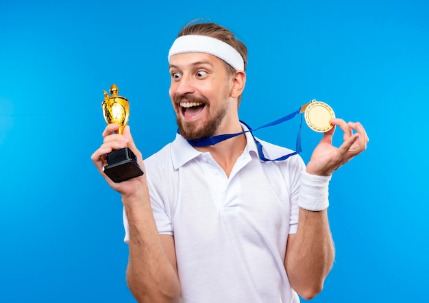 Радостный молодой красивый спортивный мужчина с повязкой на голову, браслетами и медалью на шее держит медаль и кубок победителя и смотрит на чашку, изолированную на синей стене