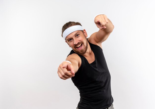 Joyful young handsome sporty man wearing headband and wristbands pointing  isolated on white wall with copy space