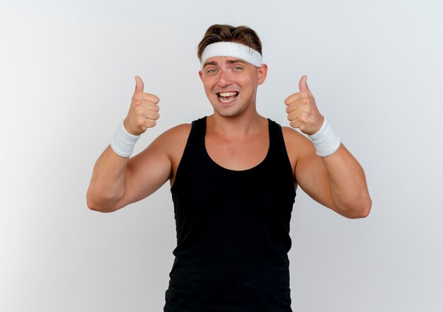 Joyful young handsome sporty man wearing headband and wristbands showing thumbs up isolated on