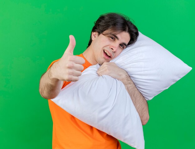 Joyful young ill man hugged pillow and showing thumb up isolated on green background