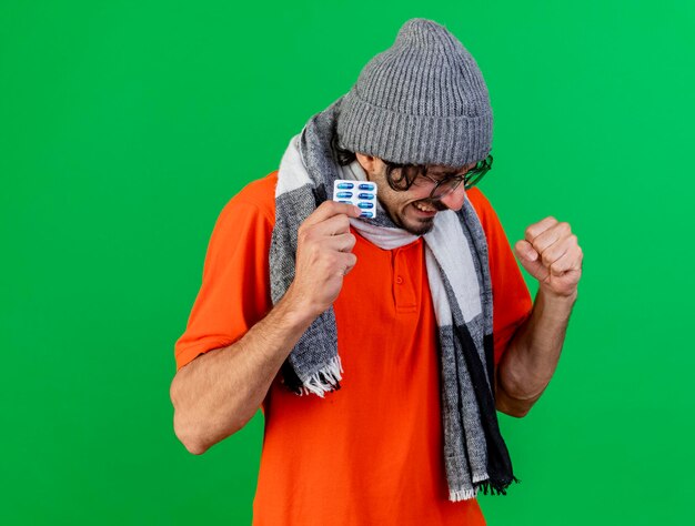 Joyful young ill man wearing glasses winter hat and scarf holding pack of medical capsules doing yes gesture isolated on green wall with copy space