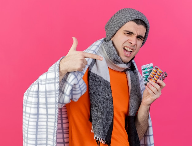 Joyful young ill man wearing winter hat with scarf wrapped in plaid holding and points at pills isolated on pink