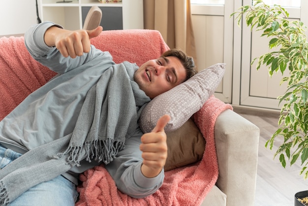 joyful young ill man with scarf around neck thumbing up lying on couch at living room