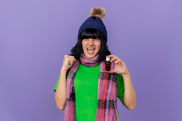 Foto gratuita gioiosa giovane donna malata che indossa cappello invernale e sciarpa che tiene il medicamento in vetro guardando il lato facendo sì gesto isolato sulla parete viola con spazio di copia