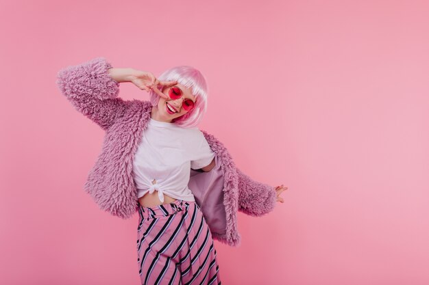 Joyful young lady in bright sunglasses smiling and dancing. Indoor photo of enchanting caucasian girl wears short peruke and trendy striped pants