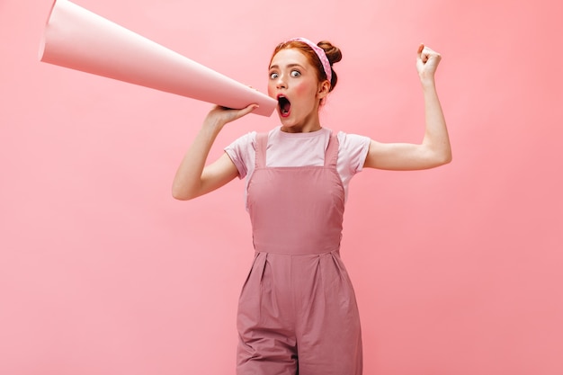 Joyful young lady in pink dress and white T-shirt screaming into mouthpiece on pink background.