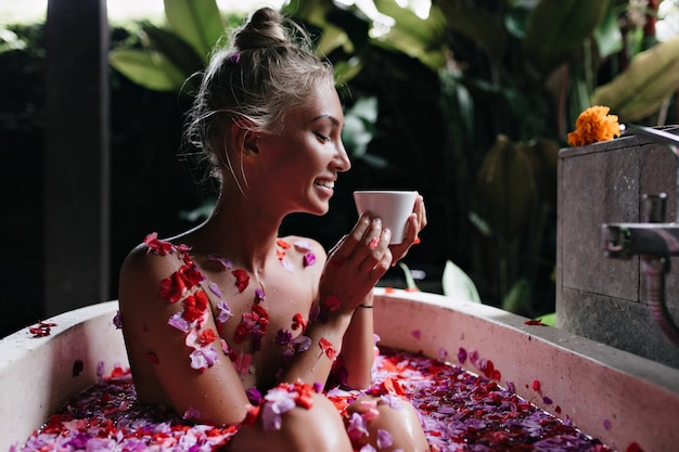 Joyful young lady relaxing during morning spa and drinking tasty tea. Winsome female model with blode hair smiling while taking bath in weekend.