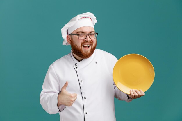 Joyful young male chef wearing glasses uniform and cap showing empty plate looking at camera showing thumb up isolated on blue background