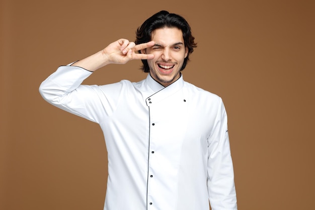 joyful young male chef wearing uniform looking at camera showing peace sign near eye winking isolated on brown background