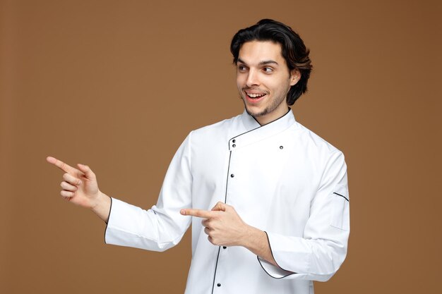 Free photo joyful young male chef wearing uniform looking at side pointing to side isolated on brown background
