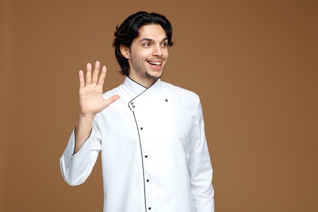 joyful young male chef wearing uniform looking at side showing hi gesture isolated on brown background