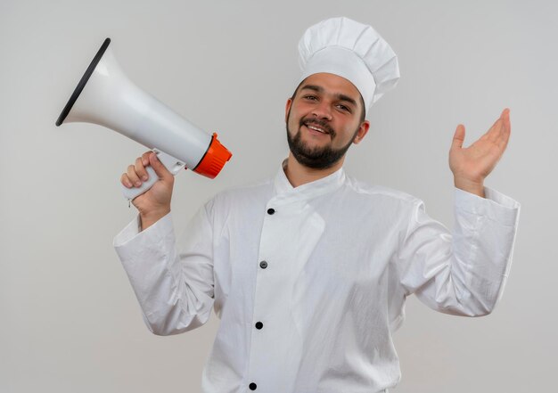 Joyful young male cook in chef uniform holding speaker and raising hand isolated on white wall