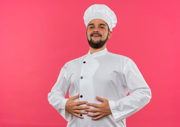 Joyful young male cook in chef uniform putting hands on belly isolated on pink wall with copy space