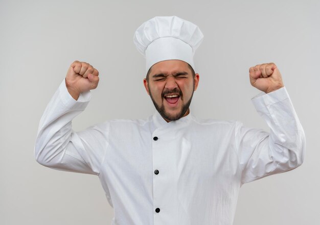 Joyful young male cook in chef uniform raising fists with closed eyes isolated on white wall