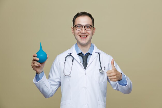 Joyful young male doctor wearing glasses medical robe and stethoscope around neck showing enema and thumb up while looking at camera isolated on purple background