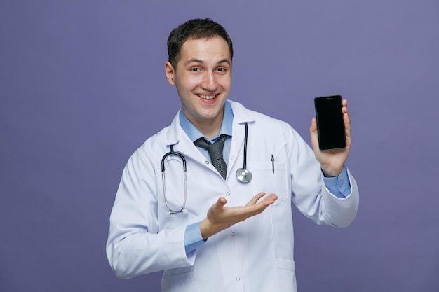 Gioioso giovane medico maschio che indossa accappatoio medico e stetoscopio intorno al collo guardando la fotocamera che mostra il telefono cellulare che punta la mano su di esso isolato su sfondo viola