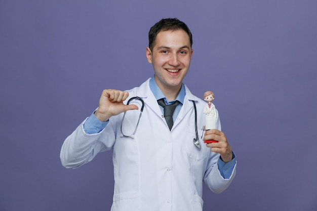 Joyful young male doctor wearing medical robe and stethoscope around neck showing doctor figurine pointing at it looking at camera isolated on purple background