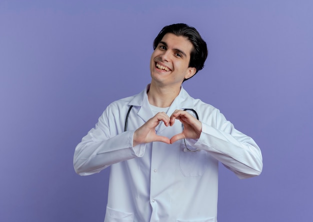Joyful young male doctor wearing medical robe and stethoscope  doing heart sign isolated on purple wall with copy space