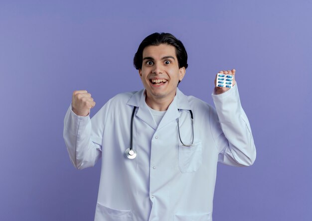 Joyful young male doctor wearing medical robe and stethoscope showing pack of capsules  doing yes gesture isolated on purple wall