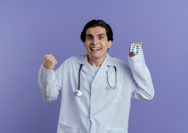 보라색 벽에 고립 예 제스처를 하 고 캡슐의 팩을 보여주는 의료 가운과 청진기를 입고 즐거운 젊은 남성 의사