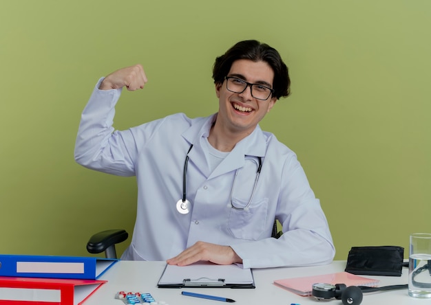 Foto gratuita gioioso giovane medico maschio che indossa veste medica e stetoscopio con gli occhiali seduto alla scrivania con strumenti medici facendo un forte gesto isolato sulla parete verde oliva