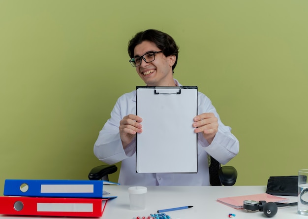 Free photo joyful young male doctor wearing medical robe and stethoscope with glasses sitting at desk with medical tools looking showing clipboard isolated