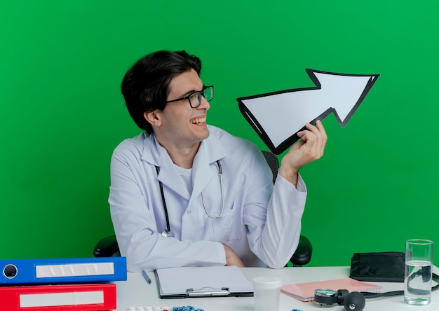 Foto gratuita gioioso giovane medico maschio che indossa veste medica e stetoscopio con gli occhiali seduto alla scrivania con strumenti medici girando la testa a lato tenendo il segno di freccia che punta sul lato isolato sulla parete verde