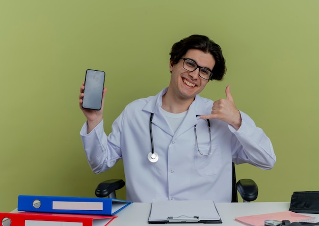 Free photo joyful young male doctor wearing medical robe and stethoscope with glasses sitting at desk