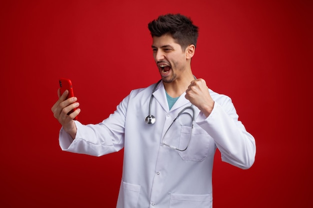 Free photo joyful young male doctor wearing medical uniform and stethoscope around his neck holding and looking at mobile phone showing yes gesture isolated on red background