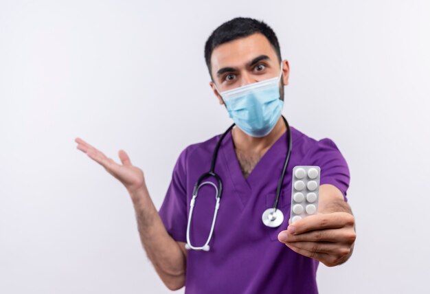 紫色の外科医の服と聴診器の医療マスクを身に着けているうれしそうな若い男性医師は、孤立した白い壁に手を上げるカメラに丸薬を差し出します
