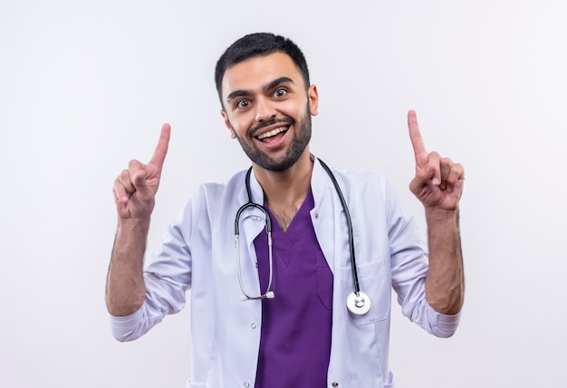 Радостный молодой мужчина-врач в медицинском халате со стетоскопом указывает вверх на изолированной белой стене