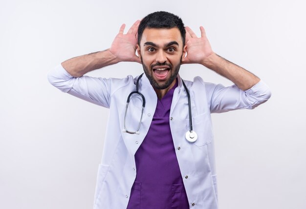 Joyful young male doctor wearing stethoscope medical gown put his hands around ears on isolated white wall