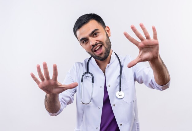 Joyful young male doctor wearing stethoscope medical gown raising hands on isolated white wall