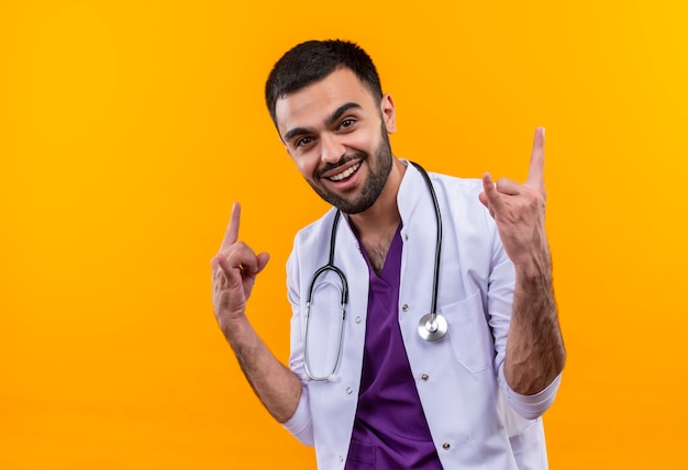Joyful young male doctor wearing stethoscope medical gown showing goat gesture on isolated yellow wall