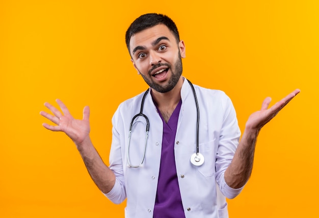 Joyful young male doctor wearing stethoscope medical gown spreads hands on isolated yellow wall