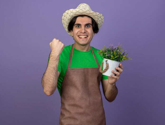 Joyful young male gardener in uniform wearing gardening hat holding flower in flowerpot showing yes gesture isolated on purple