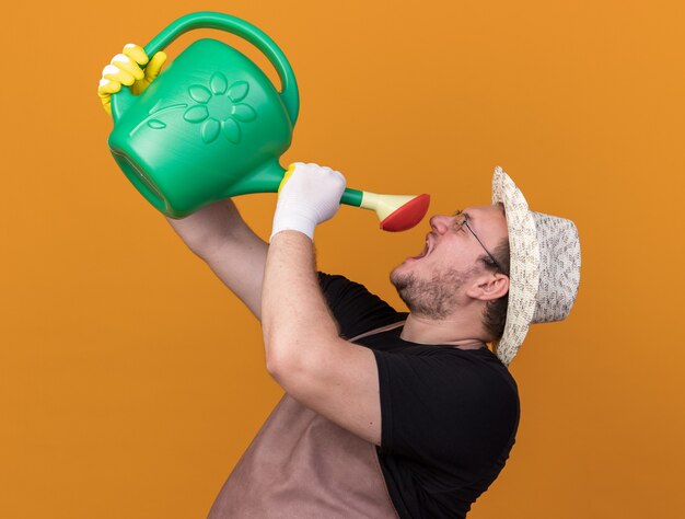 オレンジ色の壁に水まき缶で自分自身に水をまくのを保持しているガーデニング帽子と手袋を身に着けているうれしそうな若い男性の庭師