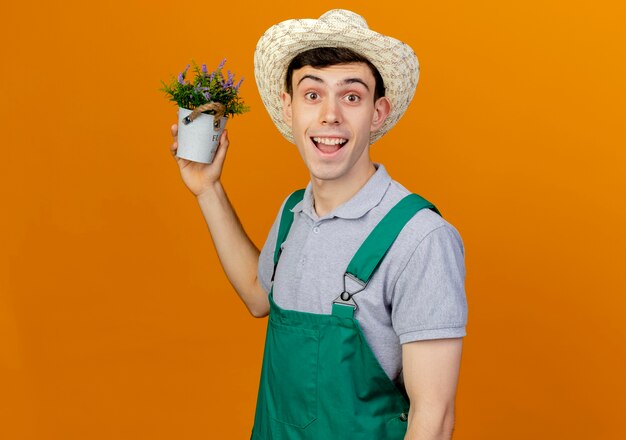 Joyful young male gardener wearing gardening hat holds flowers in flowerpot