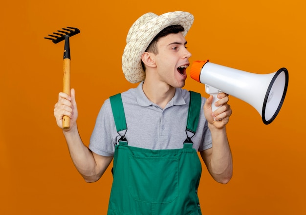 Joyful young male gardener wearing gardening hat holds rake and speaks into loud speaker looking at side 