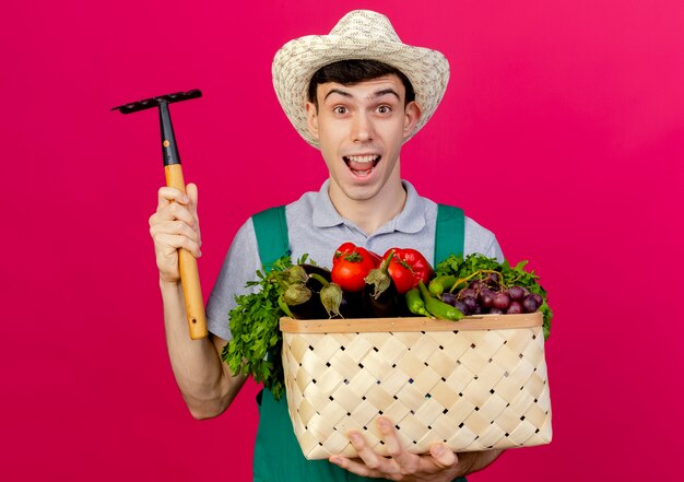 Joyful young male gardener wearing gardening hat holds vegetable basket and rake 