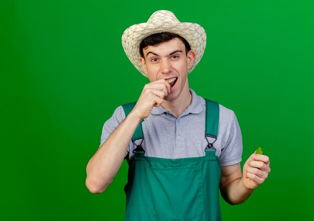 Joyful young male gardener wearing gardening hat pretends to bite broken hot pepper isolated on green background with copy space