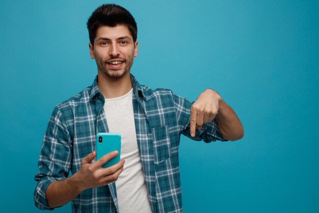 Joyful young man holding mobile phone looking at camera pointing down isolated on blue background