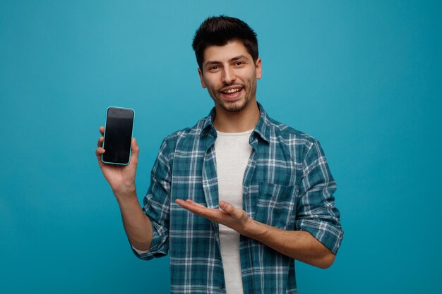 Joyful young man showing mobile phone to camera pointing at it with hand looking at camera isolated on blue background