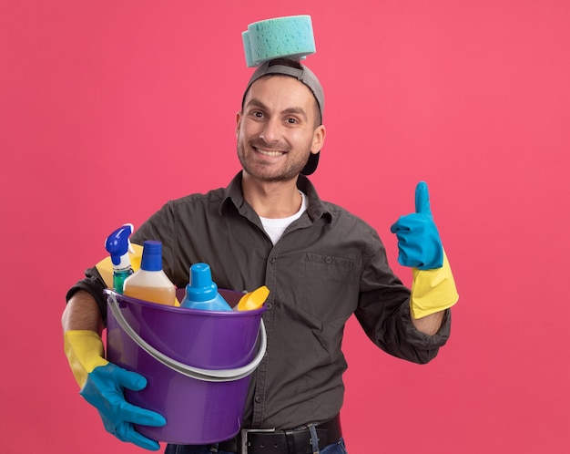 Joyful young man wearing casual clothes and cap in rubber gloves holding bucket with cleaning tools with sponge on his head  smiling showing thumbs up standing over pink wall