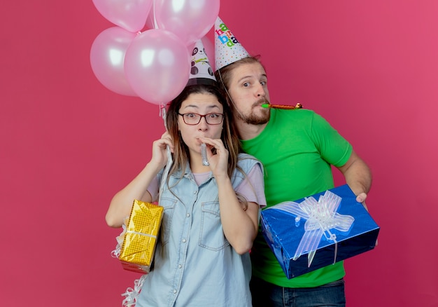 Joyful young man wearing party hat holds gift box and blows whistle standing behind surprised young girl holding helium balloons and gift box isolated on pink wall