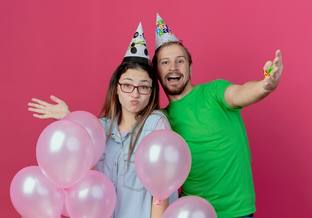 Радостный молодой человек в партийной шляпе и удивленная молодая девушка, стоящая с гелиевыми шарами, изолированными на розовой стене