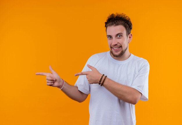 Радостный молодой человек в белой футболке указывает в сторону обеими руками на изолированной оранжевой стене