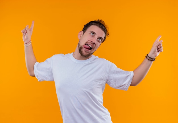 Joyful young man wearing white t-shirt showing tongue points to up on isolated orange wall