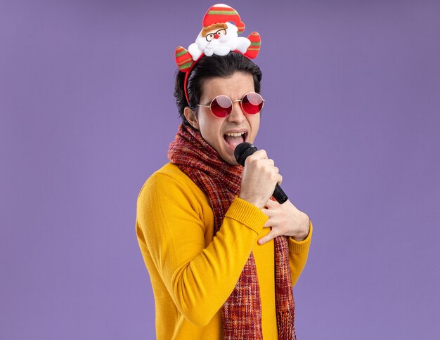 Free photo joyful young man with warm scarf around neck in yellow turtleneck and glasses with funny rim on head holding microphone singing standing over purple background